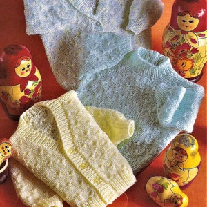 May include: Three knitted baby cardigans in blue, light blue, and yellow. The cardigans have a button closure and a cable knit pattern. The cardigans are displayed on a red background with Russian nesting dolls.