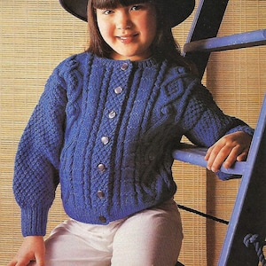 Könnte beinhalten: Ein Kind trägt einen blauen Strickcardigan mit Knopfleiste und passendem blauen Hut. Der Cardigan hat ein strukturiertes Zopfmuster. Das Kind trägt auch eine weiße Hose.