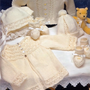 Op de afbeelding: Een set gebreide babykleding in crèmekleur, inclusief een vest, een muts, slofjes en een bonnet. De kleding ligt op een wit oppervlak met een kantversiering.