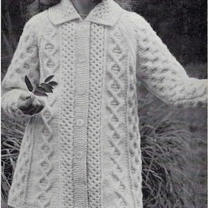 Op de afbeelding: Een jong meisje draagt een witte gebreide cardigan met een kabelbreipatroon. De cardigan heeft een knoopsluiting en een kraag.