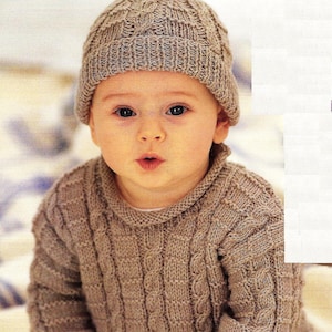 Può includere: Un bambino che indossa un maglione di lana marrone con un motivo a treccia e un cappello di lana marrone con un pon pon in cima. Il bambino è seduto su una superficie bianca e indossa jeans blu.