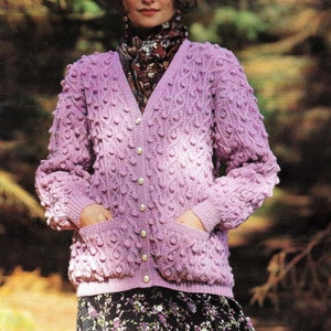 Könnte beinhalten: Ein helllila Cardigan mit strukturiertem Muster und V-Ausschnitt. Der Cardigan hat Taschen und Perlmuttknöpfe. Die Frau trägt einen gemusterten Schal und einen Blumenrock. Die Länge des Cardigans beträgt ca. 60 cm.