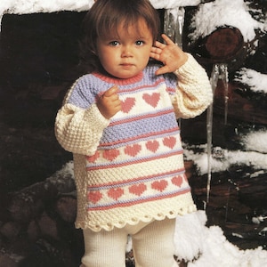 May include: A toddler wearing a white, pink, and purple knitted sweater with a heart pattern. The sweater has a white crocheted trim. The toddler is also wearing white knitted pants and red rain boots.