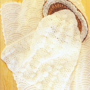 May include: A white crocheted baby blanket with a delicate lace trim. The blanket is folded and placed on a wooden surface with seashells.