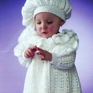 May include: A white crocheted baby dress with a ruffled collar and a matching white crocheted hat. The dress has a scalloped hem and is made with a delicate stitch pattern.