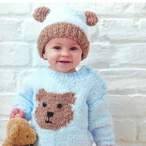 May include: A baby wearing a light blue knitted sweater with a brown bear design and a brown knitted hat with brown pom-poms. The baby is holding a brown teddy bear.