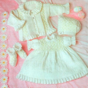 May include: A white knitted baby dress, cardigan, hat, and booties set. The dress has a delicate lace trim and a textured pattern. The cardigan has a button closure and a ruffled collar. The hat is a simple beanie style. The booties are small and have a ruffled edge.