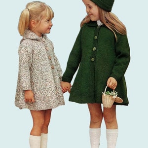 Op de afbeelding: Twee jonge meisjes dragen gebreide jassen. Eén meisje draagt een groene jas en een groene muts. Het andere meisje draagt een wit en bruin gevlekte jas. Het meisje in de groene jas houdt een klein rieten mandje met bloemen vast.