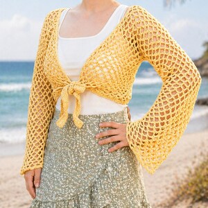 May include: A yellow crochet bolero with long sleeves tied at the front. The bolero is worn over a white vest top and paired with a green floral wrap skirt. The background shows a beach with the ocean and palm trees.