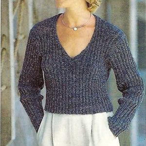 Può includere: Primo piano di una donna che indossa un maglione di lana blu e grigio con scollo a V e maniche lunghe. Indossa una gonna bianca e una collana d'argento con un ciondolo a forma di cuore.