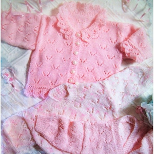 May include: Two pink knitted baby sweaters with delicate lace detailing. The top sweater has a button closure and a scalloped collar. The bottom sweater has a more open knit pattern.