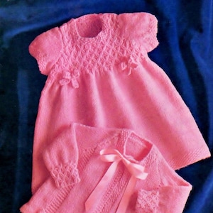 May include: A pink knitted baby dress and cardigan set. The dress has a detailed bodice and small floral accents. The cardigan features a satin ribbon tie and decorative cuffs. The set is displayed on a blue background.