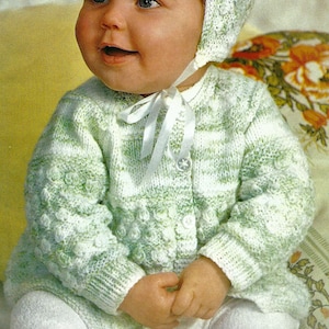May include: A baby wearing a green and white striped knitted cardigan with a matching bonnet. The cardigan has a button closure and a white ribbon tied around the neck.