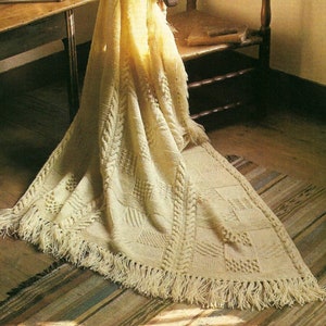 May include: A white crocheted throw blanket with fringe hangs over a wooden chair. The blanket has a cable knit pattern and is draped over the back of the chair.