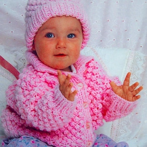 Peut inclure: Une veste de bébé en crochet rose avec un bonnet assorti. La veste a une fermeture à boutons et un design texturé. Le bébé porte un pantalon à fleurs violet et blanc.