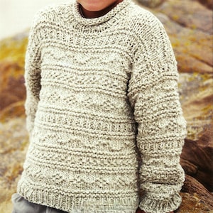 Könnte beinhalten: Ein junger Junge trägt einen hellbeigen Strickpullover mit einem strukturierten Muster. Der Pullover hat einen runden Halsausschnitt und lange Ärmel.