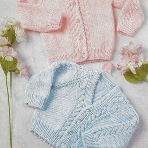 Może przedstawiać: Dwa dzianinowe kardigany dla niemowląt, jeden różowy, a drugi jasnoniebieski, z wzorem warkocza i zapięciem na guziki.