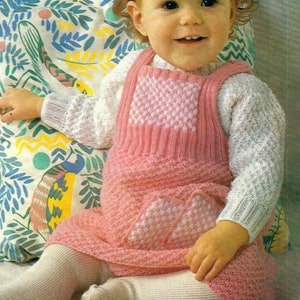 May include: A young child wearing a pink knitted dress with white accents. The dress has a checkered pattern on the front and a pocket on the side. The child is sitting on a colourful patterned cushion.