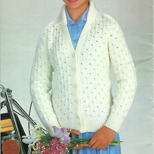 May include: A white knitted cardigan with a button-up front and a delicate openwork pattern. The cardigan has a collar and long sleeves. The model is wearing a light blue shirt underneath.