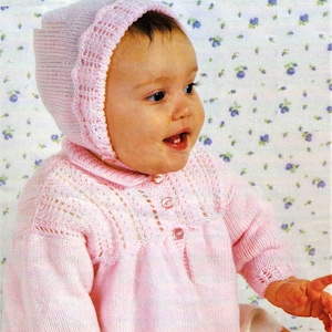 May include: A pink knitted baby cardigan with a matching bonnet. The cardigan has a button closure and a delicate lace pattern. The baby is wearing a white undershirt.