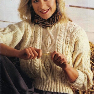 May include: A woman wearing a cream-colored cable knit sweater with a black and white patterned scarf. She is holding a knitting needle in her hands.