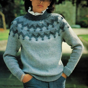 May include: A blue and grey knitted sweater with a geometric pattern on the neckline. The sweater has long sleeves and a round neckline. The model is wearing a black hat.