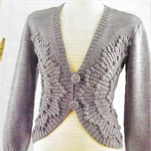 Op de afbeelding: Een grijze gebreide cardigan met een V-hals en een knoopsluiting. De cardigan heeft een textuurontwerp met verhoogde bulten aan de voorkant.