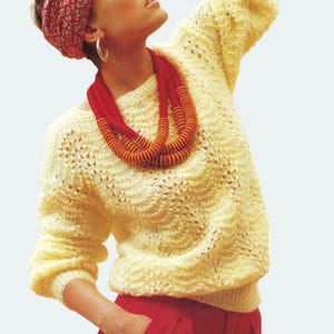 May include: A pale yellow knitted jumper with a round neck and long sleeves. The jumper features a textured, open-weave pattern. A red and orange beaded necklace and a red patterned headband complete the outfit. The model is also wearing red trousers.