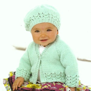 May include: A baby wearing a light green knitted cardigan and matching beret. The cardigan has a delicate lace pattern around the edges. The baby is sitting on a colorful patterned skirt.
