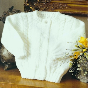 Könnte beinhalten: Ein weißer gestrickter Baby-Cardigan mit Knopfleiste und Zopfmuster.