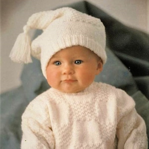 Könnte beinhalten: Ein Baby in einem cremefarbenen Strickpullover und einer passenden Nachtmütze mit Quaste. Der Pullover hat ein strukturiertes Muster, und die Mütze hat eine lange, spitze Form. Das Baby sitzt und schaut in die Kamera.