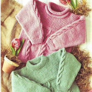 May include: Two knitted sweaters, one pink and one green, with a cable knit pattern. The sweaters are folded and displayed on a burlap background.