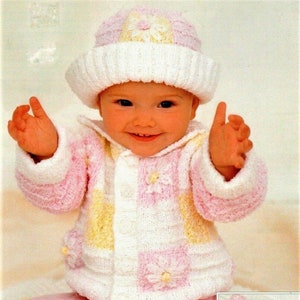 Puede incluir: Un bebé con un cárdigan de punto rosa y amarillo con un gorro de punto blanco. El cárdigan tiene un patrón de margaritas y el gorro tiene una flor en la parte superior. El bebé está sentado en una manta rosa y amarilla.