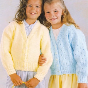 May include: Two young girls wearing pastel-colored knitted cardigans. One cardigan is pale yellow, the other is light blue. Both cardigans have button closures. The girls are smiling and wearing skirts. One girl is holding a basket.