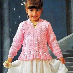 May include: A pink knitted cardigan with a cable knit pattern and a white bow detail. The cardigan has a V-neck and long sleeves. The model is wearing a white skirt and a pearl necklace.
