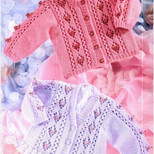 May include: Two knitted baby cardigans with a delicate lace pattern and pink flower embellishments. The cardigan on top is pink and the cardigan on the bottom is lavender.