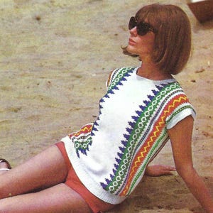 May include: A white knit top with a colorful geometric pattern in green, blue, yellow, and red. The top has short sleeves and is paired with red shorts. The person is wearing sunglasses and is lying on the sand.