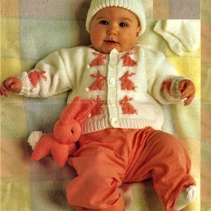 May include: A white knitted baby cardigan with pink bunny appliques, worn by a baby wearing a white knitted hat and pink pants. The baby is holding a pink knitted bunny toy.