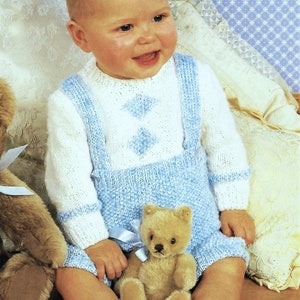 May include: A baby wearing a white and blue knitted sweater with a matching blue knitted overall. The baby is sitting on a white and blue patterned surface and holding a teddy bear.