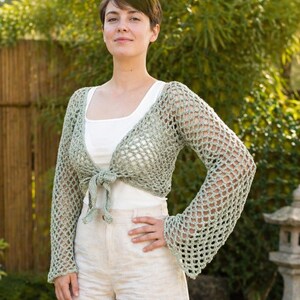 May include: A pale green, open-weave crochet shrug with long sleeves and a tie closure. The shrug is worn over a white top and paired with beige shorts. The crochet design features a net-like pattern, offering a delicate and airy aesthetic.