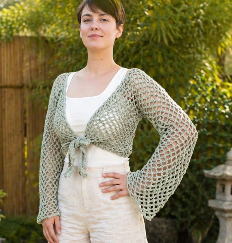 May include: A pale green, open-weave crochet shrug with long sleeves and a tie closure. The shrug is worn over a white top and paired with beige shorts. The crochet design features a net-like pattern, offering a delicate and airy aesthetic.