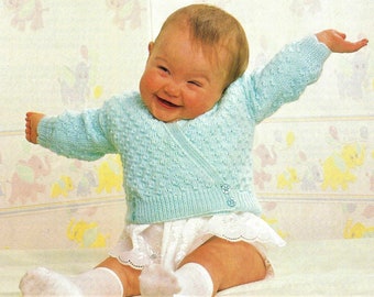 May include: A baby wearing a light blue knitted cardigan with a button closure. The cardigan has a textured pattern and is worn over a white lace-trimmed undershirt.