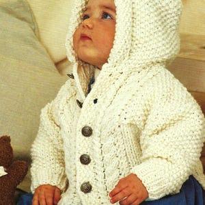 May include: A white knitted baby cardigan with a hood and button fastening. The cardigan features a cable knit pattern and is being worn by a baby.