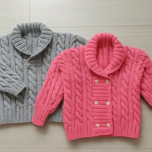 May include: Two cable-knit cardigans. One is grey, the other is pink. Both have shawl collars and button fastenings. The sweaters are made of a thick, textured yarn, with a prominent cable knit pattern.