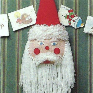 May include: A white yarn Santa Claus head with a red hat and a white pom-pom on top. The Santa Claus head is hanging from a brown ring. The background is a green and brown striped wallpaper.