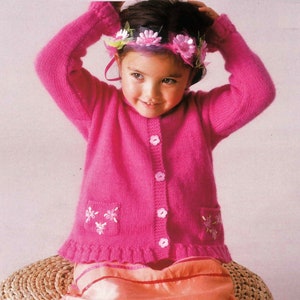 May include: A pink knitted cardigan with a frilled hem and embroidered flowers. The cardigan has a button fastening and pockets on the front.