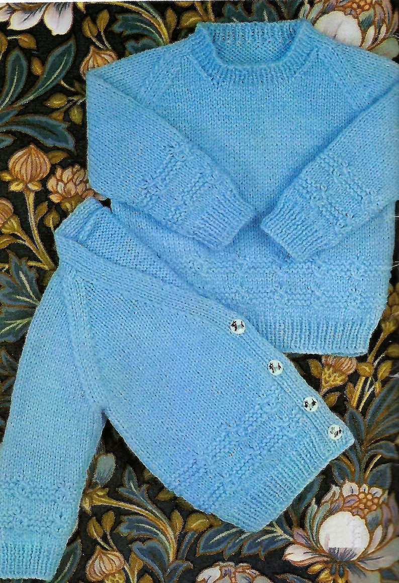 May include: Two light blue knitted sweaters, one with buttons, laid out on a floral patterned fabric.