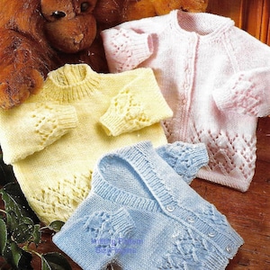 Może przedstawiać: Trzy dziergane kardigany dla niemowląt w różnych kolorach: żółtym, różowym i niebieskim. Każdy kardigan ma delikatny wzór koronki i zapięcia na guziki.
