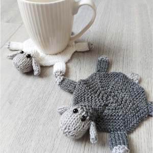 Może przedstawiać: Dwa dzianinowe podkładki pod kubki w kształcie owiec, jeden biały, a drugi szary, z czarnymi oczami. Biała podkładka znajduje się pod białym ceramicznym kubkiem z uchwytem.