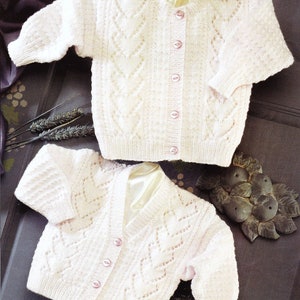 May include: Two white knitted baby cardigans with a delicate lace pattern and pink buttons. The cardigans are laid out on a dark background.